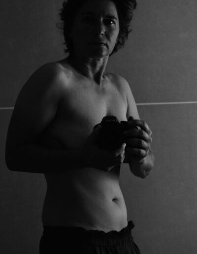 Black and white self-portrait of a photographer holding a Nikon camera against a tiled wall in soft low light