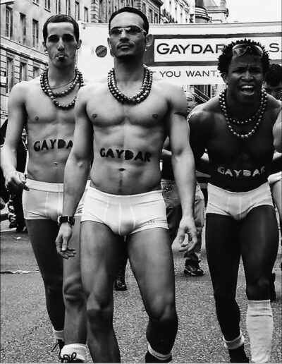 Black and white street photograph of three men in white briefs and bead necklaces marching in a Pride parade