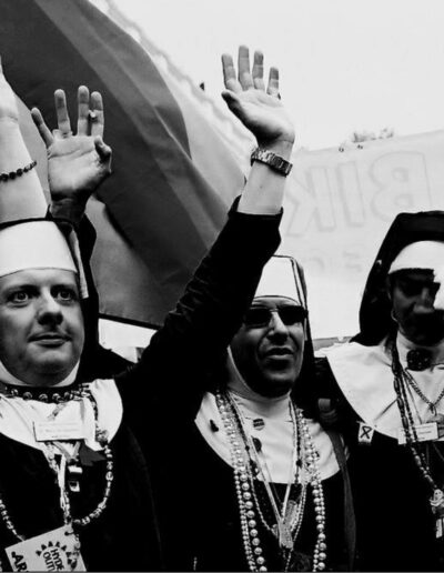 Black and white photograph of people dressed as nuns raising their hands in celebration beneath a rainbow flag at Pride