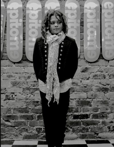 Black and white portrait of a young person with dreadlocks, a buttoned jacket and a floral scarf against a brick wall of skateboards