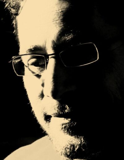 Sepia high-contrast close-up portrait of a bearded man in glasses, half his face in deep shadow