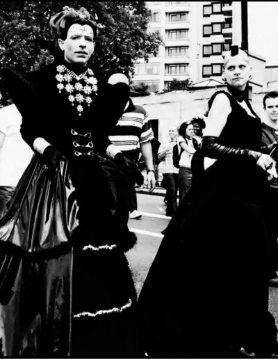 Black and white street photograph of two people in dramatic gothic gowns and jewellery at a Pride event