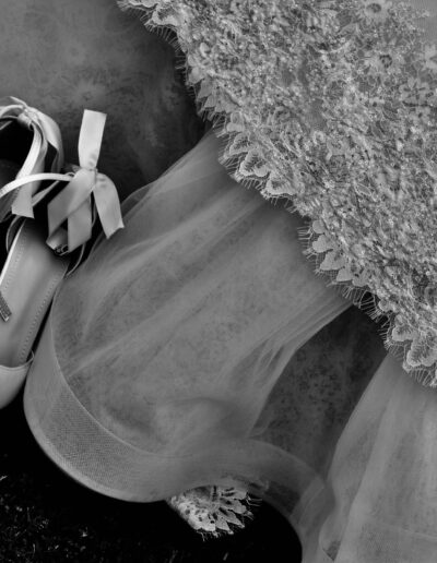 Black and white photograph of ivory bridal shoes with satin bows resting on a lace and tulle wedding gown on the grass