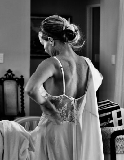 Black and white photograph of a bride from behind in a lace-backed slip dress, hair in a low bun, getting ready at home