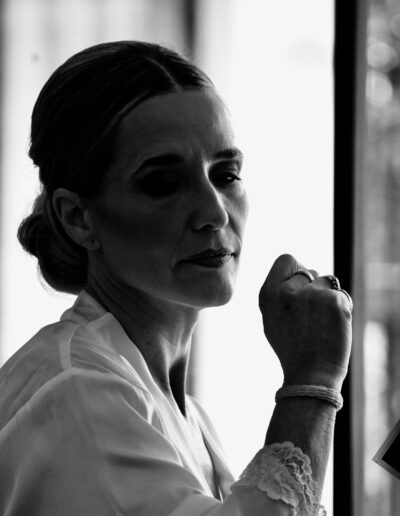 Black and white backlit portrait of a bride in a lace-trimmed robe, lit softly by a window while getting ready