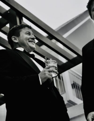 Black and white low-angle photograph of three smiling groomsmen in tuxedos holding whisky glasses under a pergola