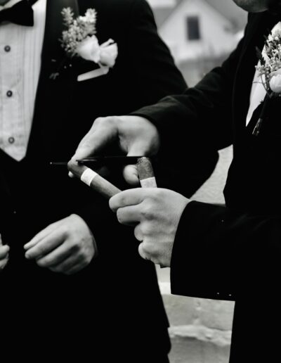 Black and white close-up of groomsmen in tuxedos and buttonholes cutting a cigar before the wedding