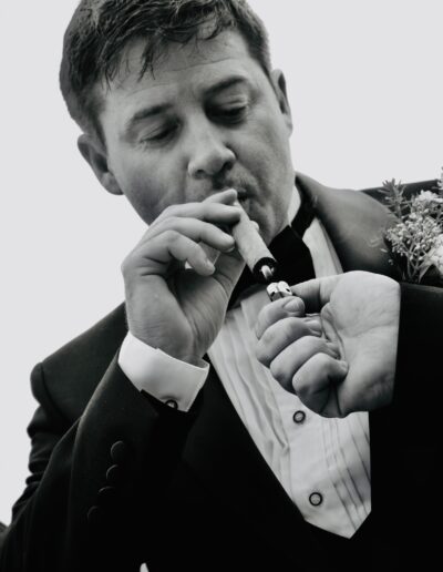 Black and white portrait of a groom in a tuxedo lighting a cigar, a buttonhole on his lapel