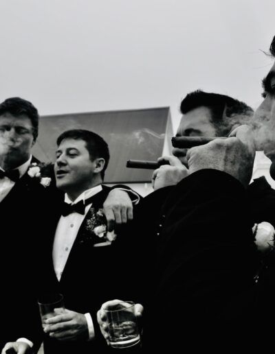 Black and white photograph of groomsmen in tuxedos sharing cigars and drinks on a rooftop terrace