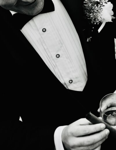 Black and white detail of a man in a pleated tuxedo shirt and bow tie holding a cigar and a glass of whisky